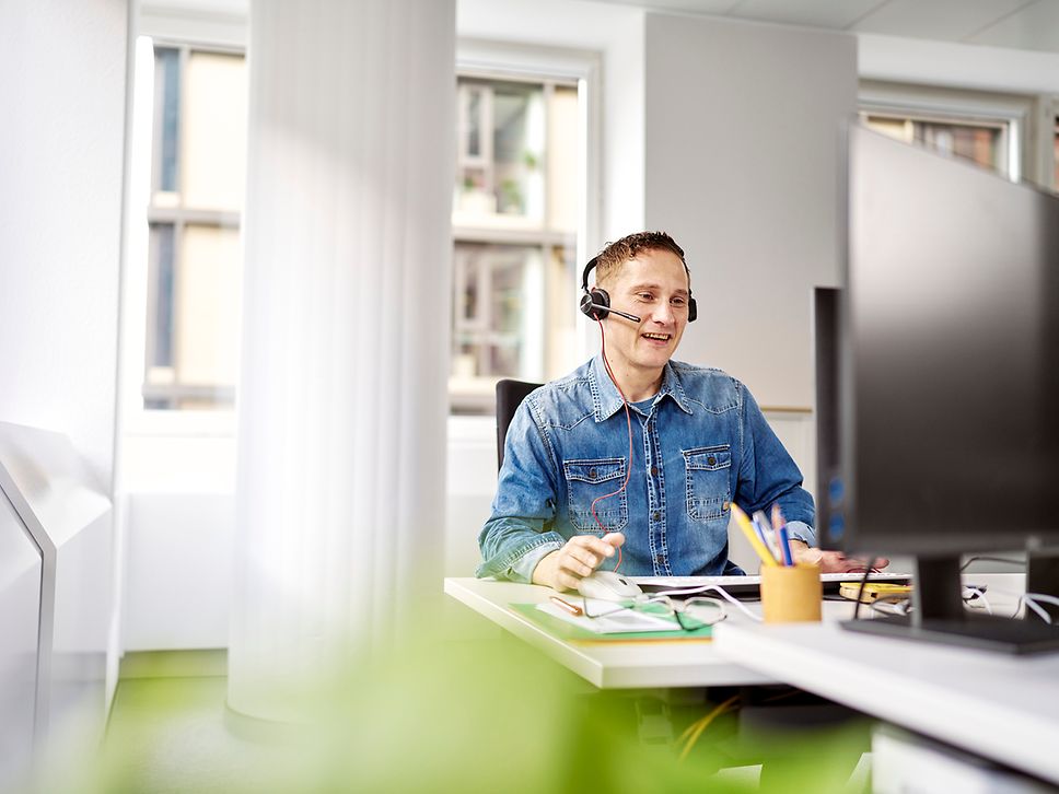 Ein Mann mit Headset sitzt an seinem Arbeitsplatz vor dem Rechner und spricht.