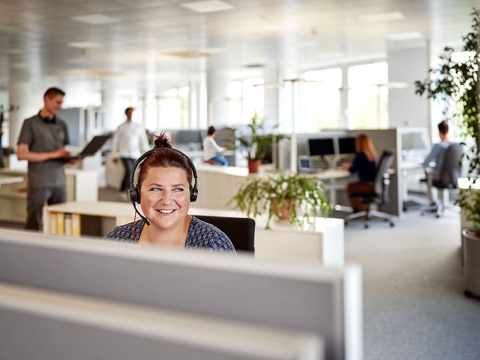 Eine Frau lächelt vor dem Rechner und trägt ein Headset