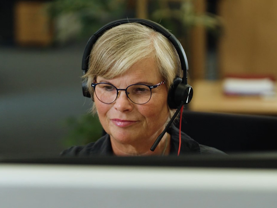 Frau sitzt lächelnd vor dem Rechner und hat ein Headset aufgesetzt
